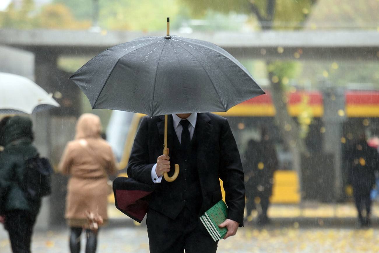 Na zdjęciu jesień w Warszawie. Mężczyzna z parasolem za jego plecami ludzie w kurtkach przeciwdeszczowych.