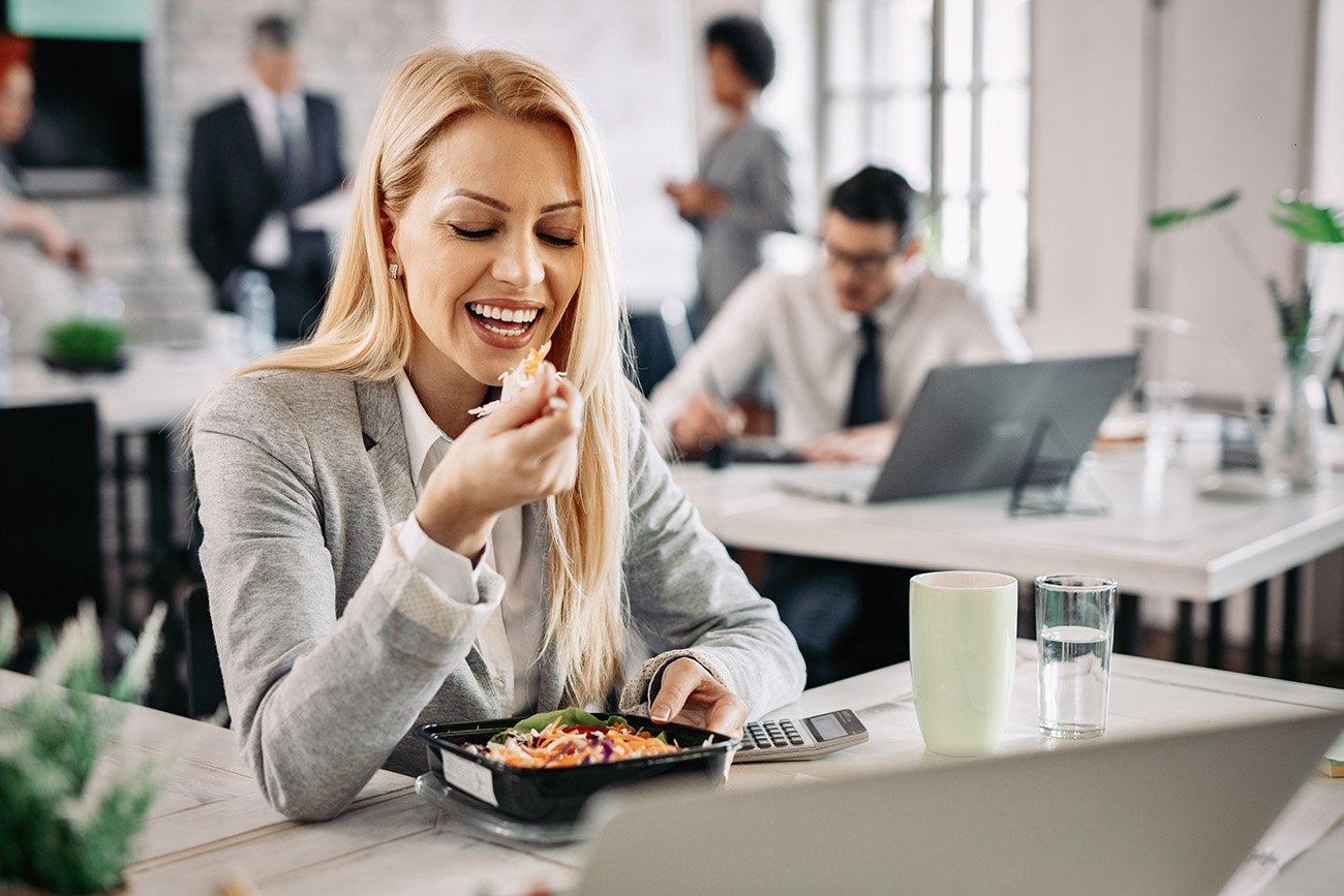 Kobieta jedząca lunch z pudełka przy biurku w pracy - tytułowa.