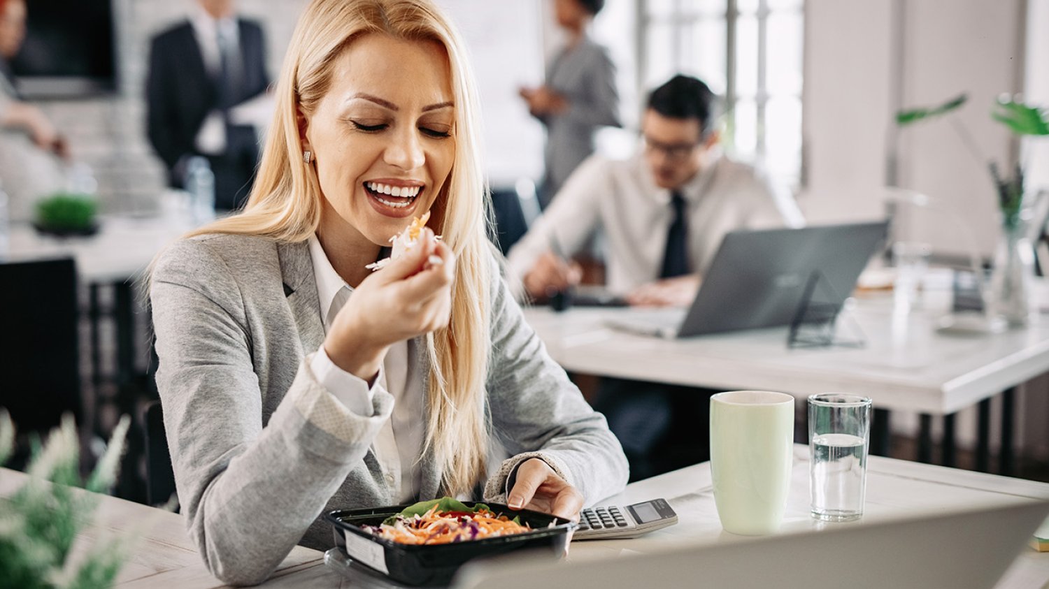 Kobieta jedząca lunch z pudełka przy biurku w pracy - tytułowa.