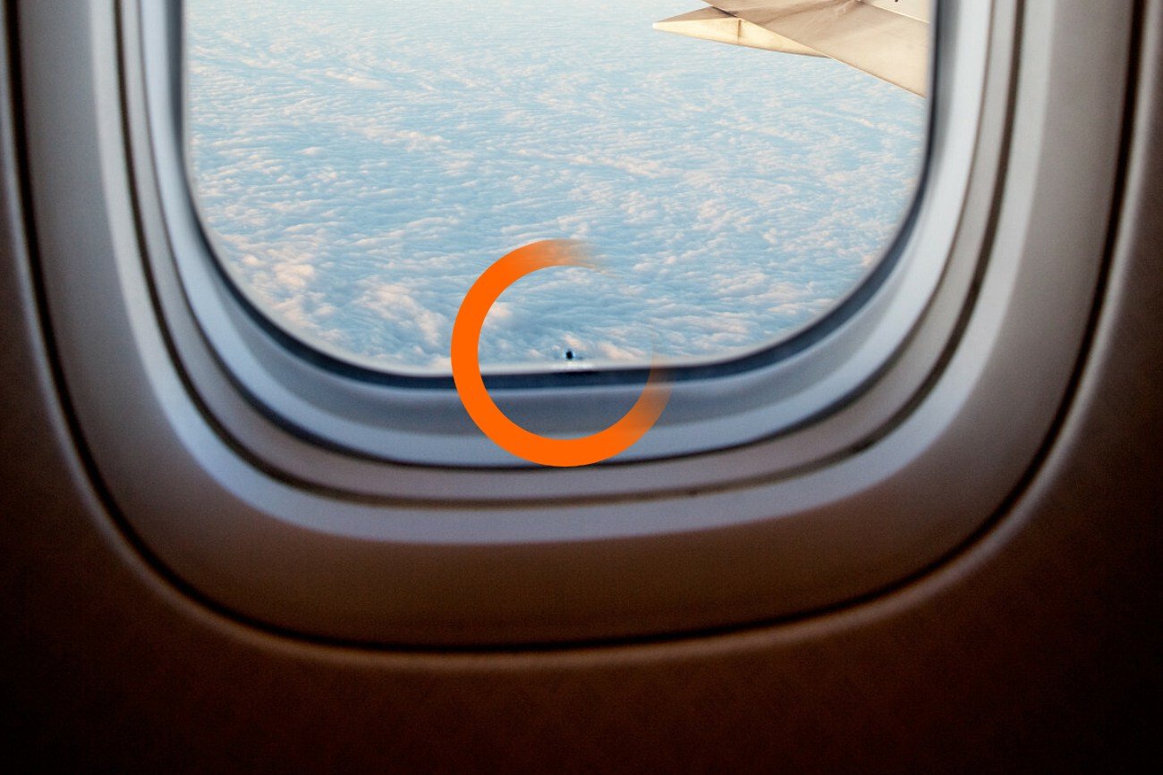 Otwór w szybie samolotu. Mała dziurka nawiercona w środkowej warstwie okna statku powietrznego. W tle chmury i fragment skrzydła.