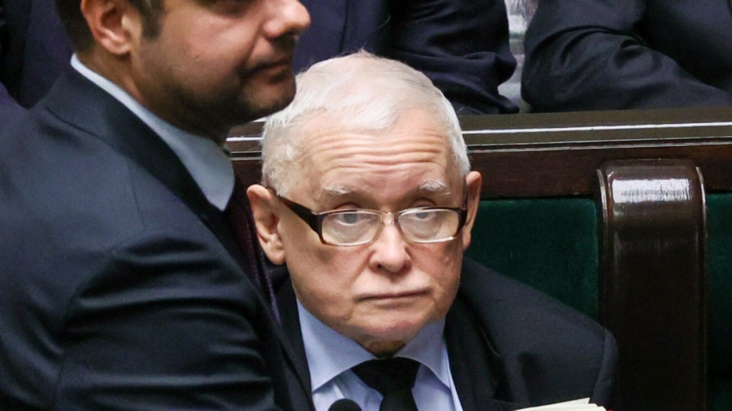 Jarosław Kaczyński siedzi w ławach sejmowych z książką w ręku. Obok na stojąco rzecznik PiS Rafał Bochenek.