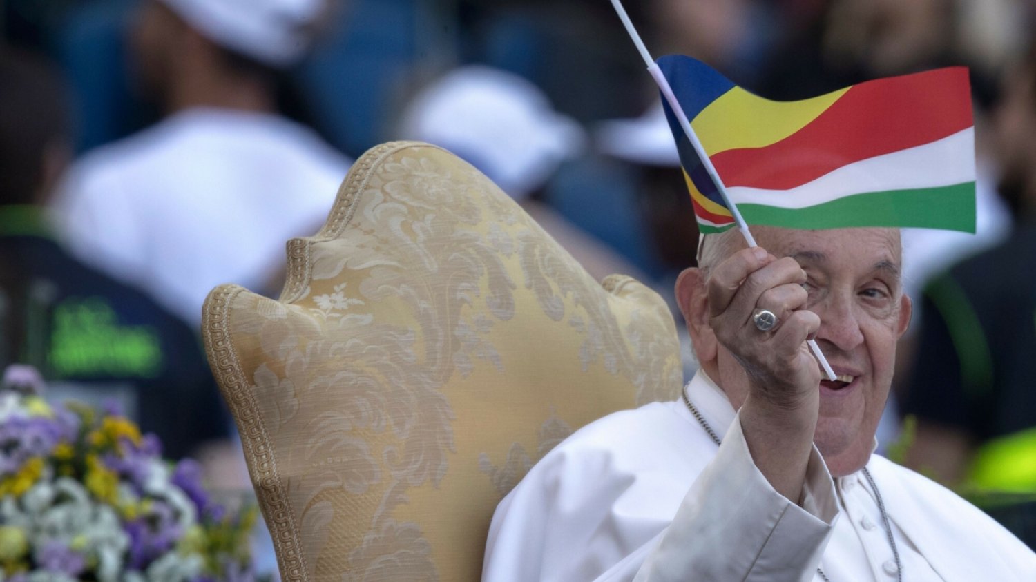 Papież Franciszek siedzi na krześle, uśmiecha się i macha flagą.