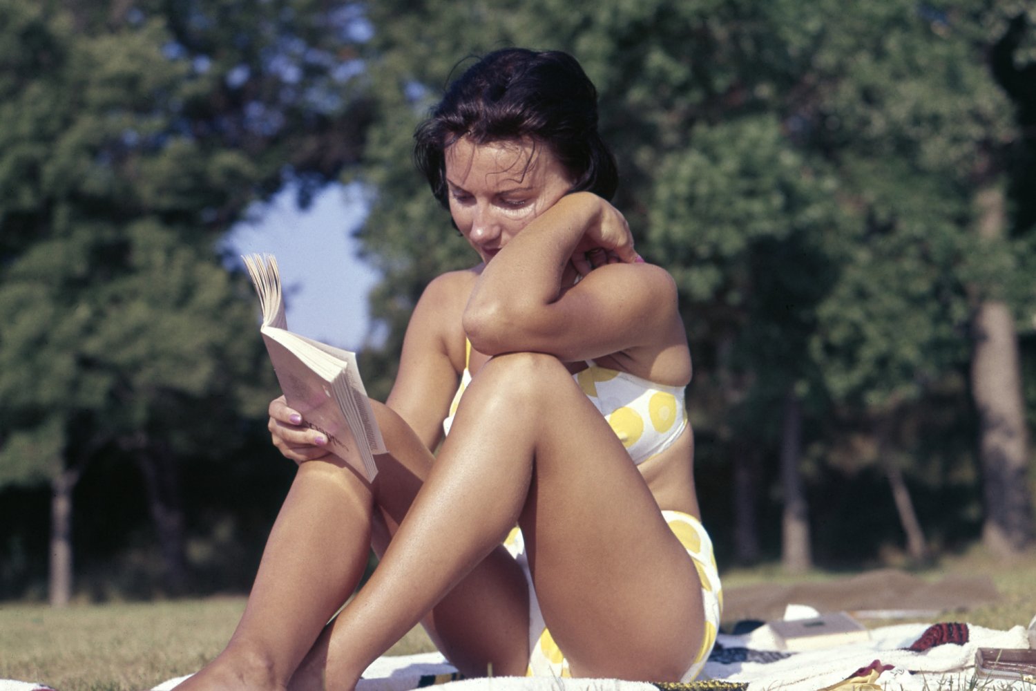 kobieta w kostiumie kąpielowym czytająca książkę na kocu na trawie