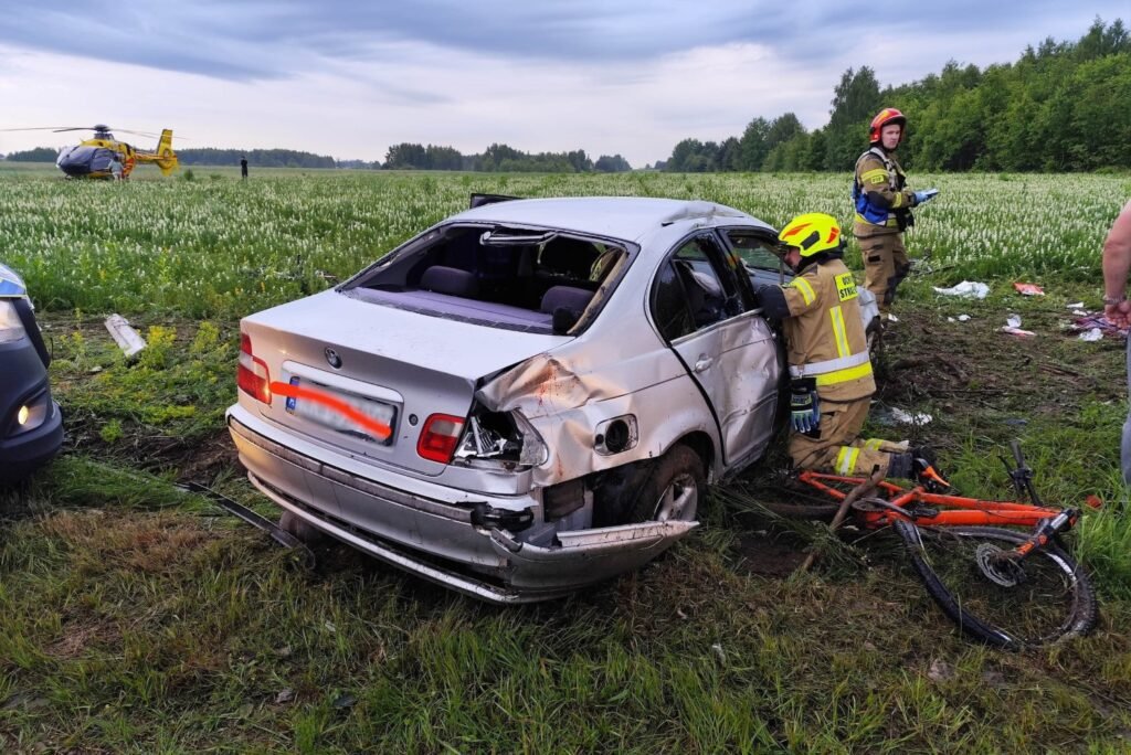 Wypadek w Końskich. Kierowca BMW wjechał autem w rowerzystów. Samochód na polu po wypadku. Wgnieciona karoseria, potłuczone szkło. Przy prawych przednich drzwiach auta klęczy strażak.