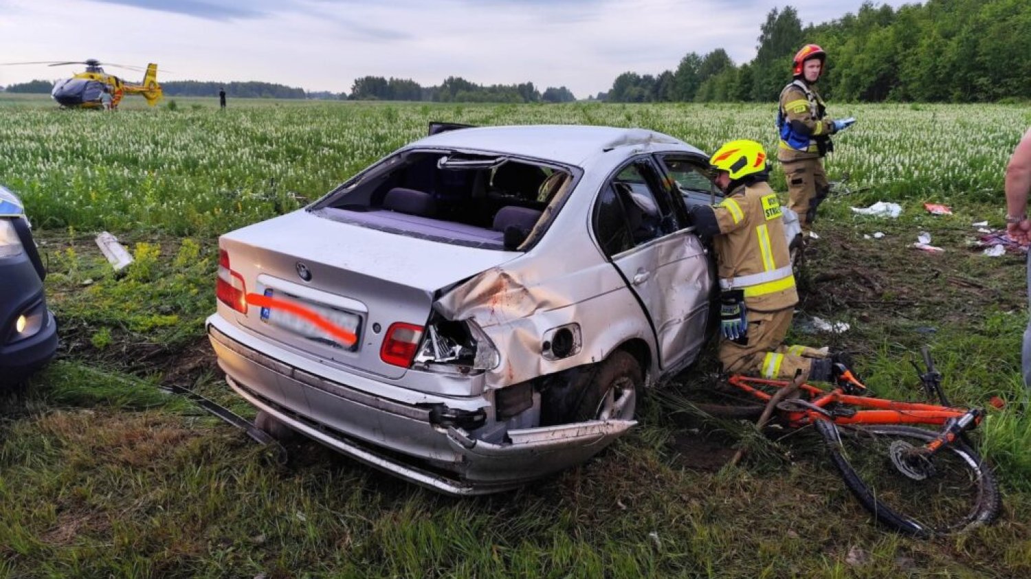 Wypadek w Końskich. Kierowca BMW wjechał autem w rowerzystów. Samochód na polu po wypadku. Wgnieciona karoseria, potłuczone szkło. Przy prawych przednich drzwiach auta klęczy strażak.