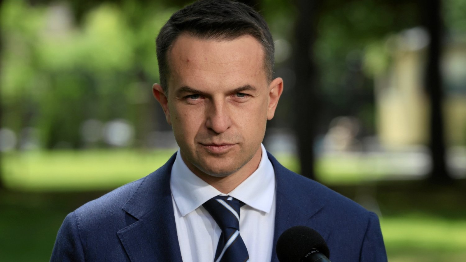 Adam Szłapka w garniturze i przy mikrofonie na tle drzew.