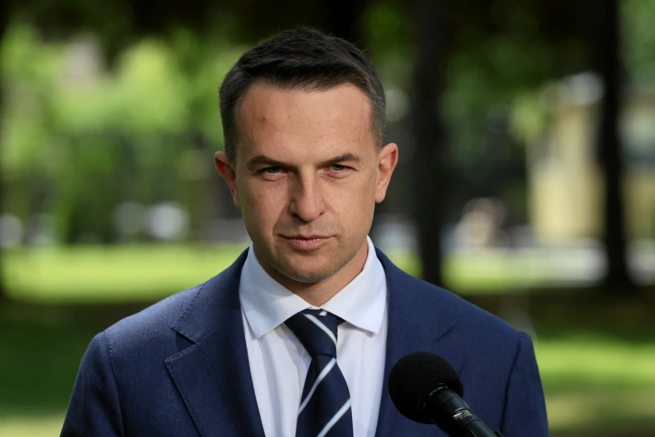 Adam Szłapka w garniturze i przy mikrofonie na tle drzew.