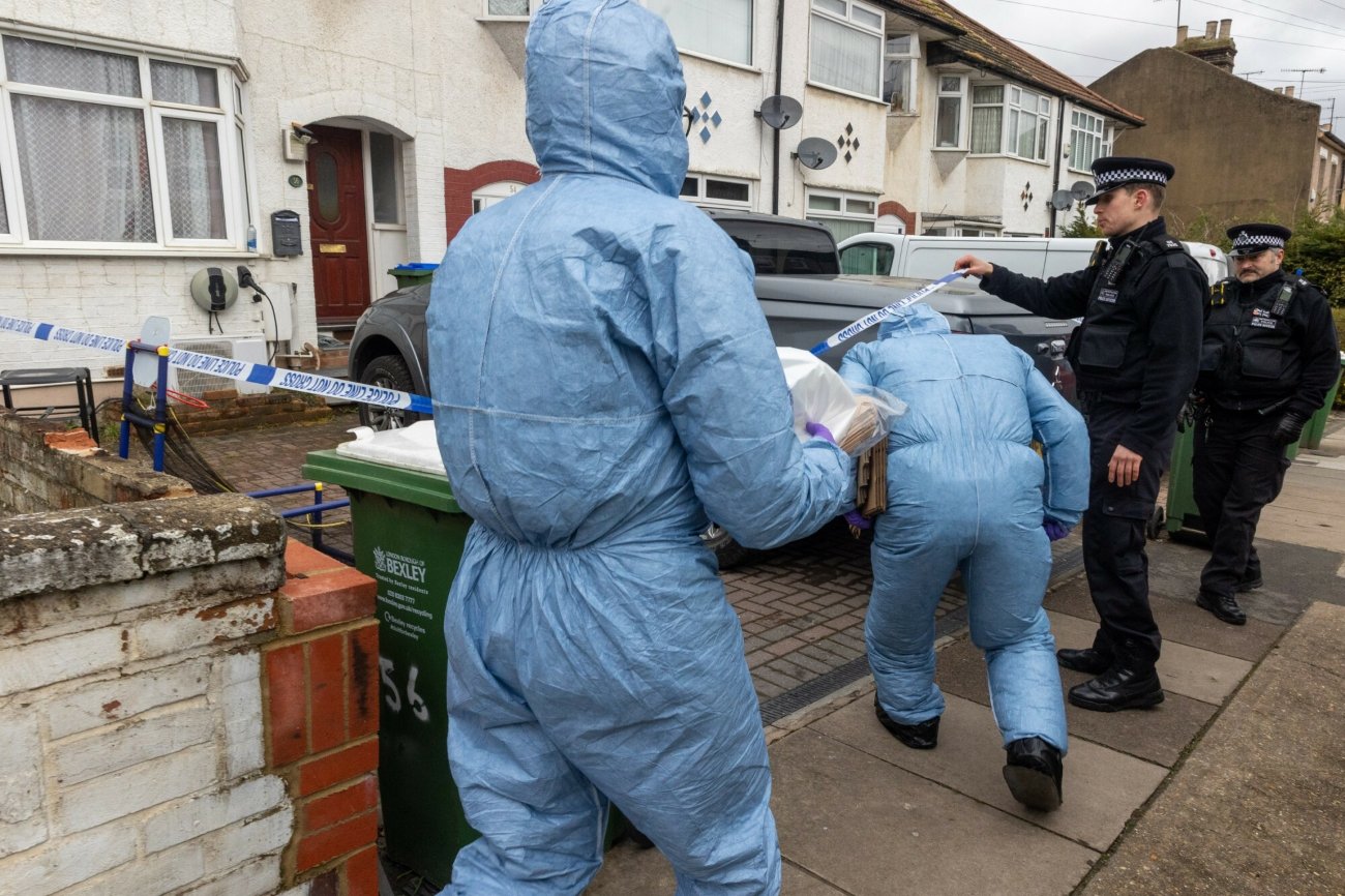 Technicy w kombinezonach ochronnych przechodzący przez taśmę z napisem "policja" do domu, w którym doszło do tragedii. Obok brytyjscy policjanci.