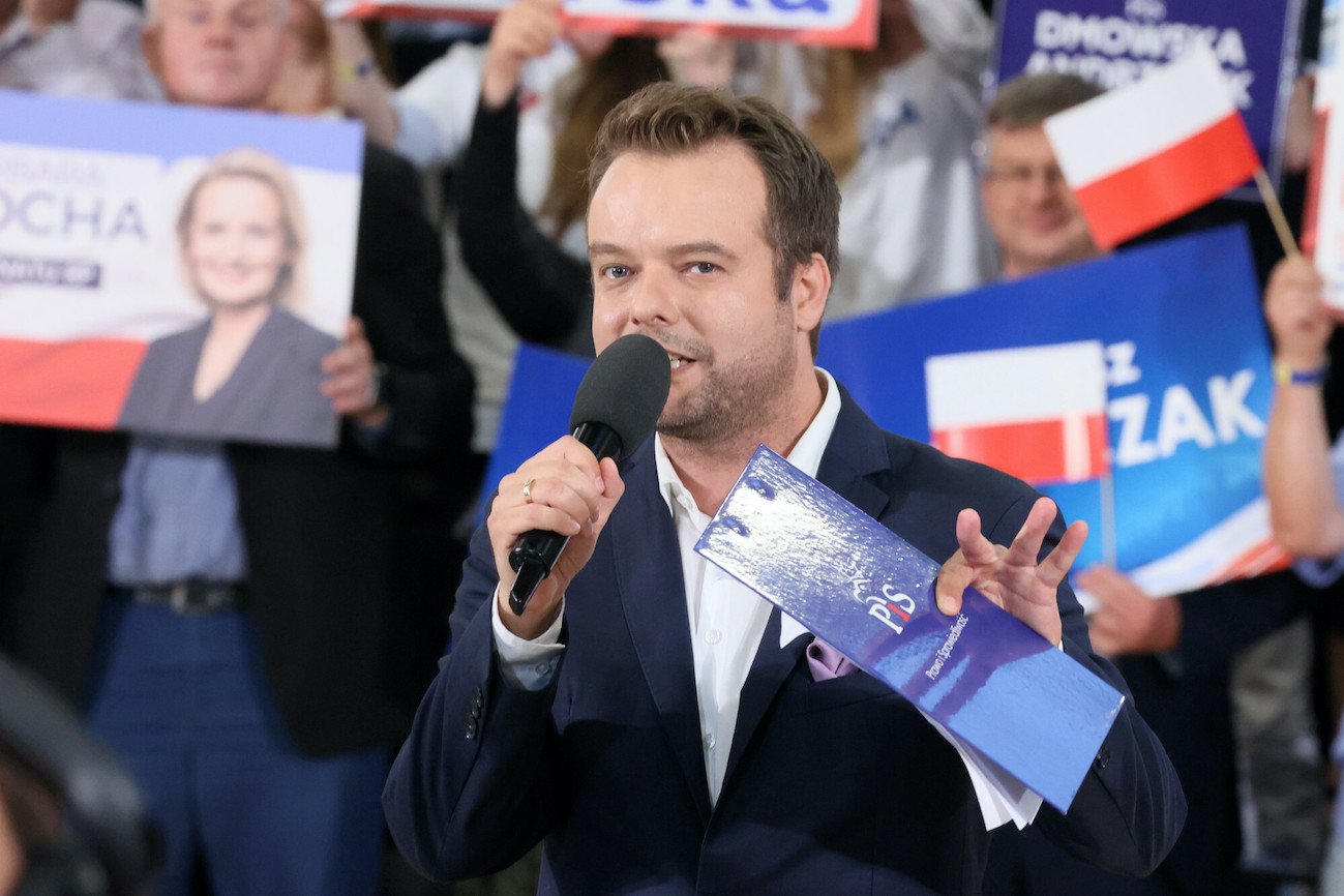 Rafał Bochenek, rzecznik prasowy Prawa i Sprawiedliwości, na konwencji wyborczej z podkładką z logo PiS w otoczeniu polityków obozu władzy.