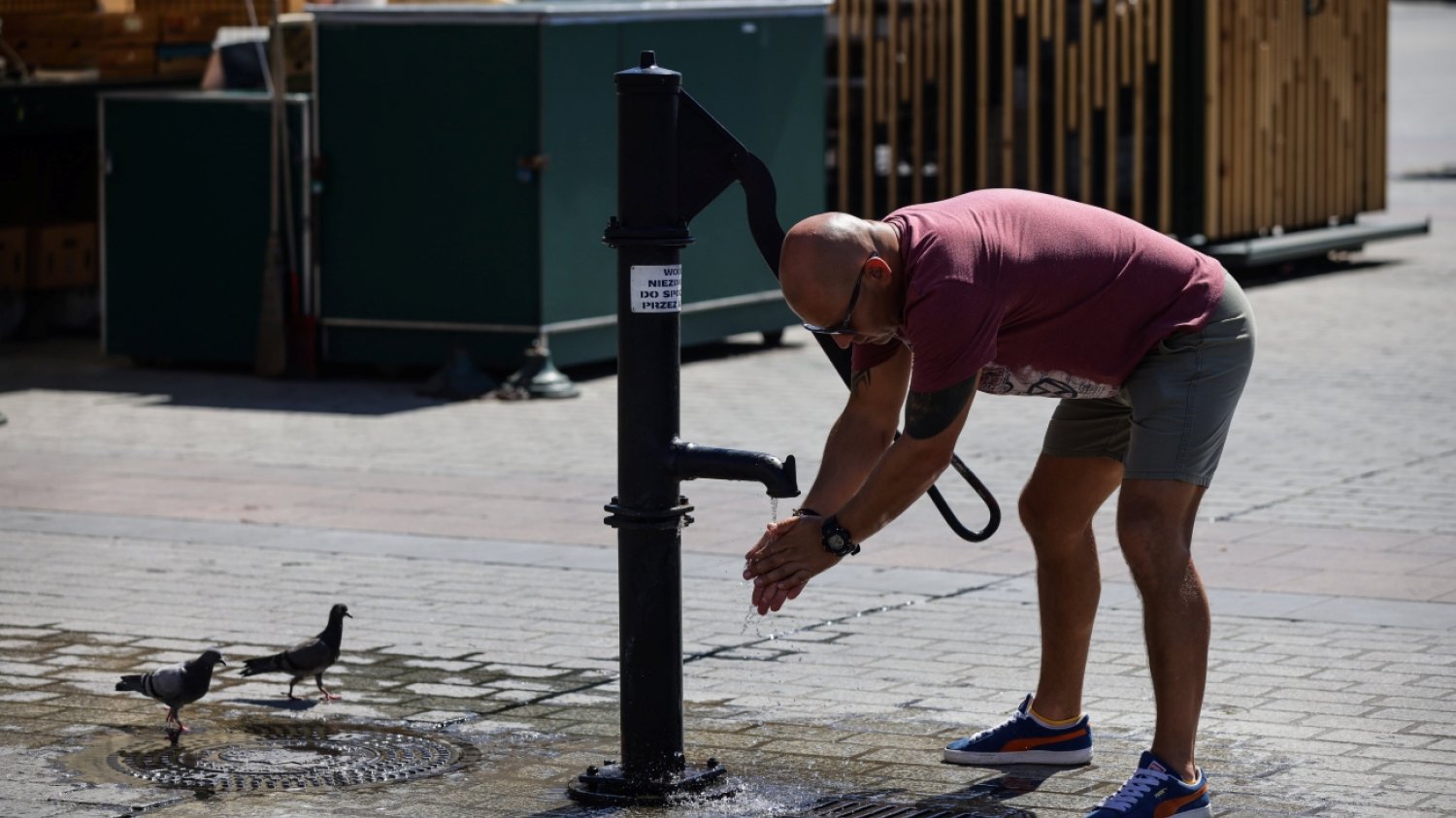 Kraków, Rynek Główny, słoneczny dzień.. Mężczyzna w t-shircie i krótkich spodenkach schyla się do wody płynącej z publicznego kranu. Obok gołębie stojące na mokrym chodniku.