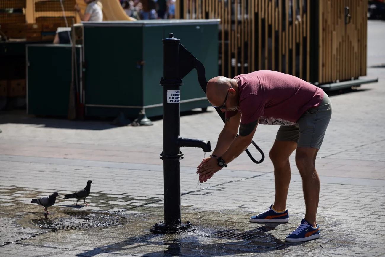 Pogoda na trzy dni będzie kapryśna. IMGW przewiduje, że wyda alerty z kilku różnych powodów. Chodzi o ostrzeżenia przed burzą, upałem i wiatrem. Kraków, Rynek Główny, słoneczny dzień.. Mężczyzna w t-shircie i krótkich spodenkach schyla się do wody płynącej z publicznego kranu. Obok gołębie stojące na mokrym chodniku.