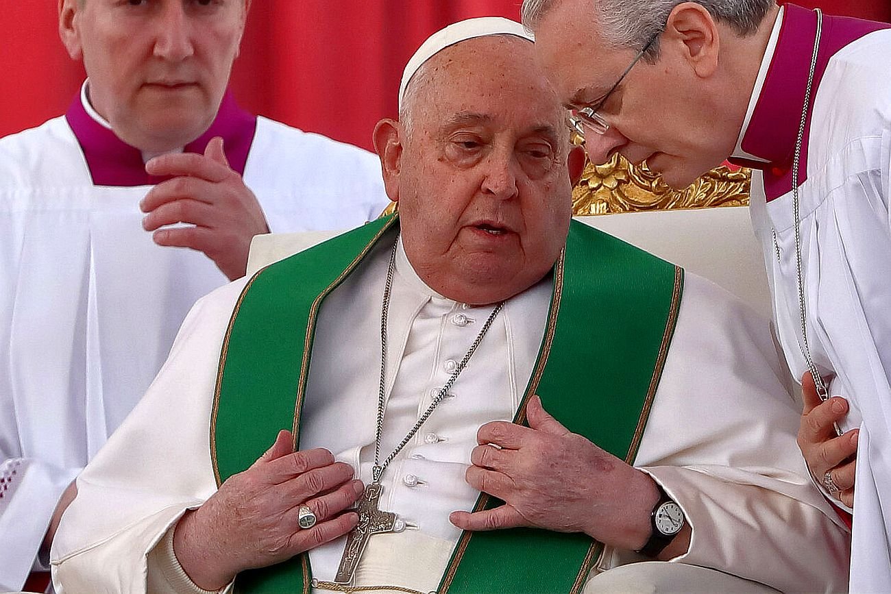 Papież Franciszek siedzi z poważną miną. Pochylają się nad nim dwaj księża.