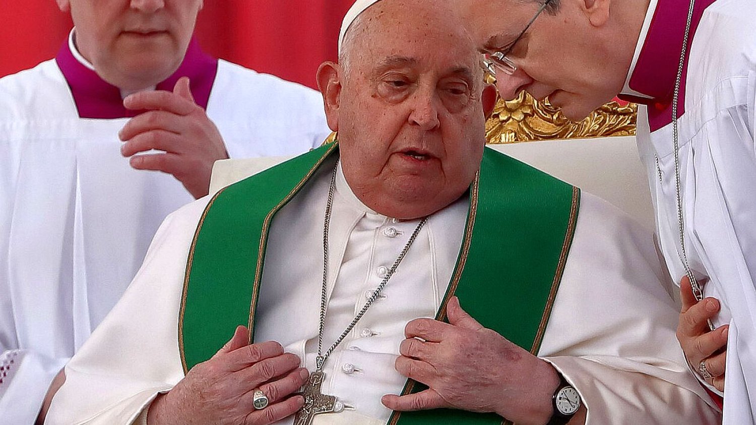Papież Franciszek siedzi z poważną miną. Pochylają się nad nim dwaj księża.