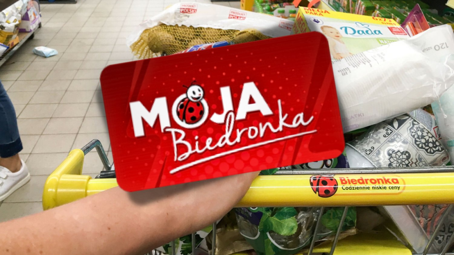 Karta Moja Biedronka pozwala skorzystać z ciekawych promocji przed 11 listopada. Na zdjęciu Karta Moja Biedronka, a w tle koszyk zakupów w sklepie Biedronka.