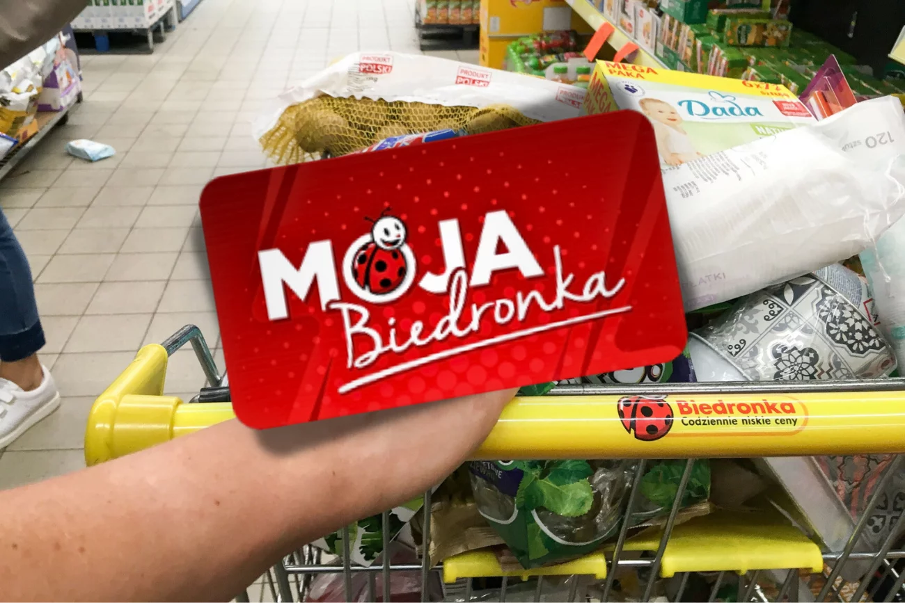 Karta Moja Biedronka pozwala skorzystać z ciekawych promocji przed 11 listopada. Na zdjęciu Karta Moja Biedronka, a w tle koszyk zakupów w sklepie Biedronka.