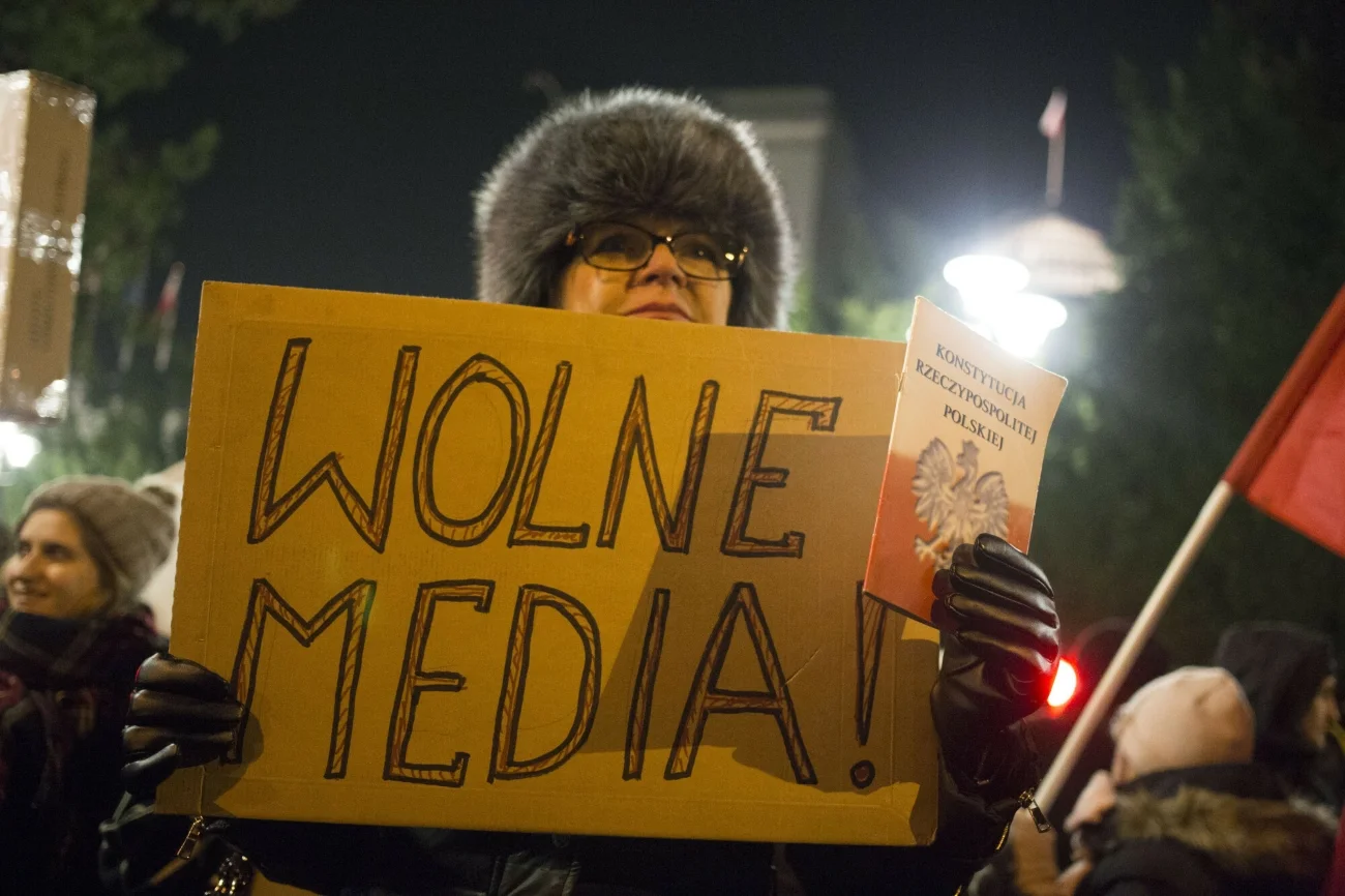 Kobieta ubrana w kurtkę i czapkę z transparentem w ręce z napisem "Wolne Media". W drugiej dłoni trzyma Konstytucję Rzeczypospolitej Polskiej.