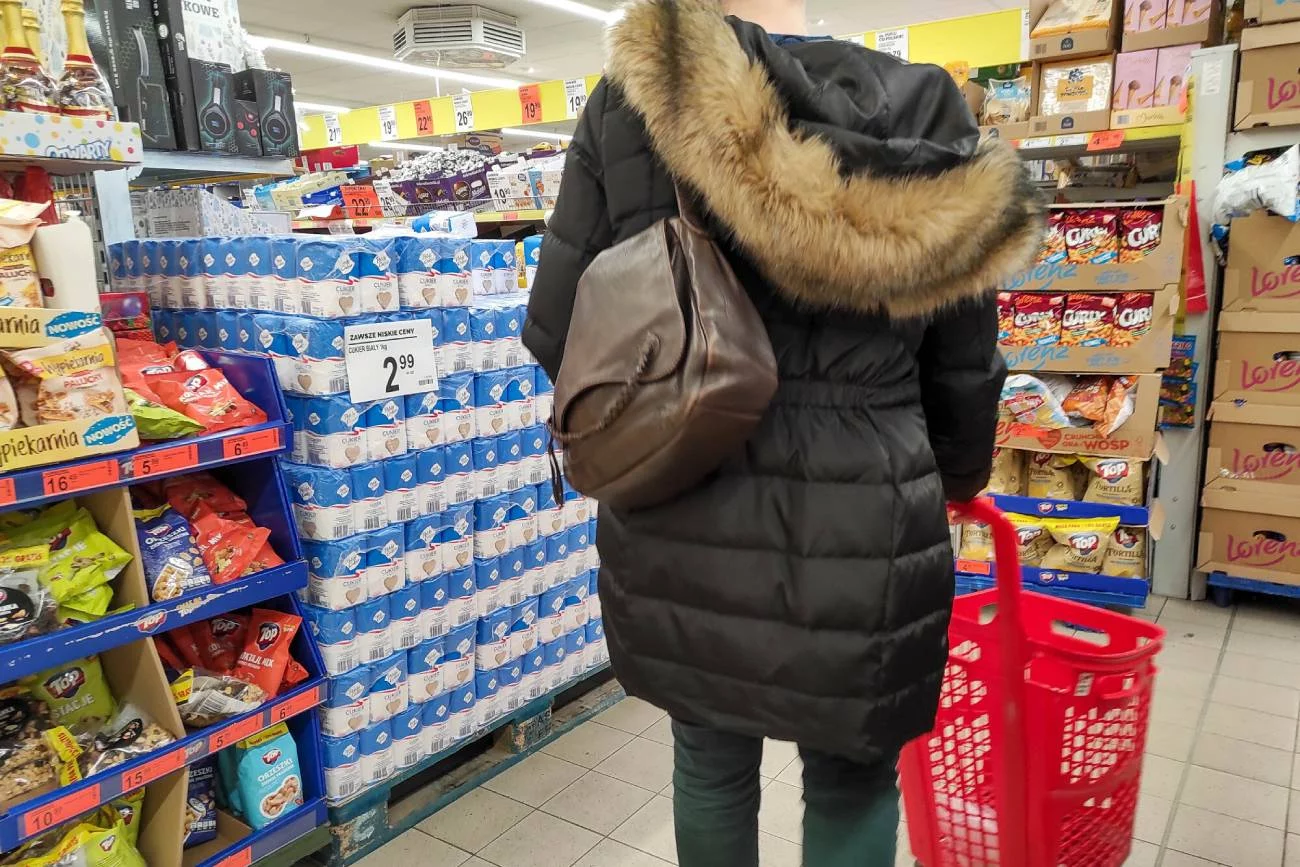 Osoba pomiędzy półkami w sklepie Biedronka. Sklep robi promocję na cukier.