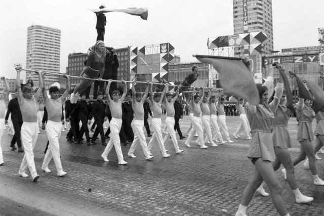 Gimnastycy niosący drążki z ćwiczącymi gimnastyczkami podczas przemarszu ul. Marszałkowską. W tle widoczne budynki "Ściany Wschodniej".