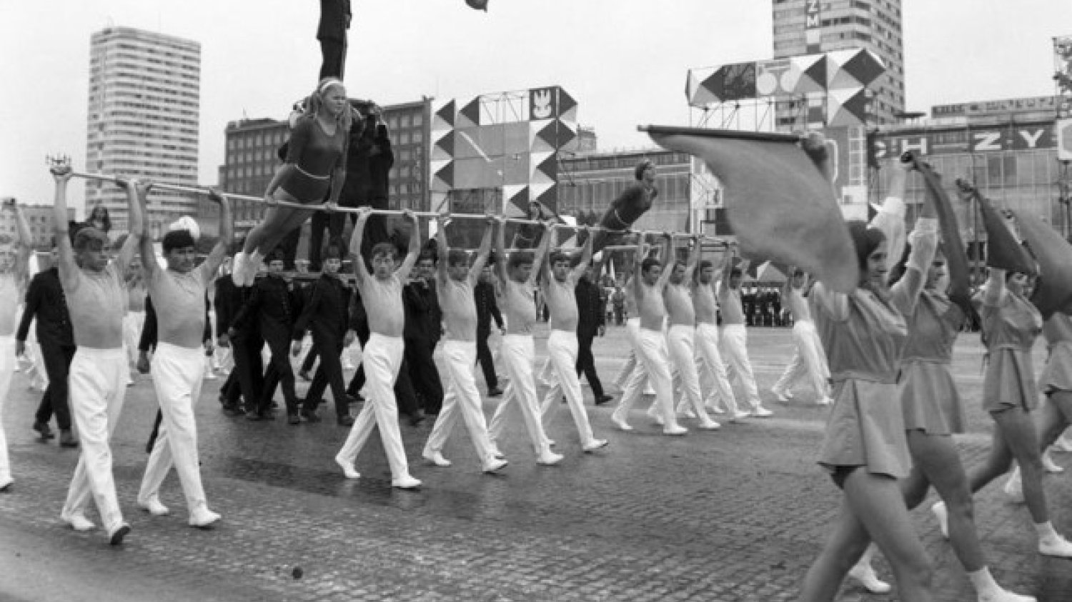 Gimnastycy niosący drążki z ćwiczącymi gimnastyczkami podczas przemarszu ul. Marszałkowską. W tle widoczne budynki "Ściany Wschodniej".
