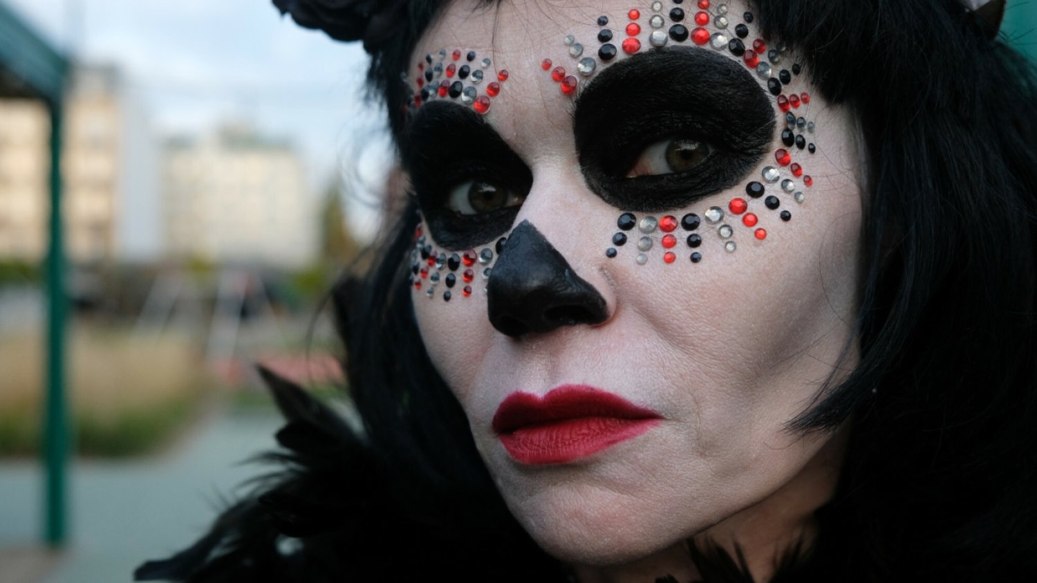 Spooky Bazar-Halloween na Nowym Bazarze Różyckiego Kobieta przebrana z okazji Halloween. Usta ma pomalowane czerwoną szminką, na szyi ma czarne boa. Wokół oczu ma naklejone cekiny, a jej skóra twarzy jest pomalowana na czarno i biało.