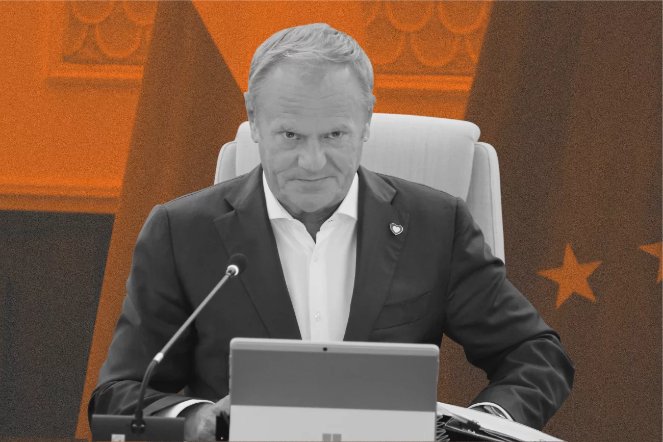Donald Tusk siedzący przy stole przy mikrofonie. Za nim flaga Polski i Unii Europejskiej.
