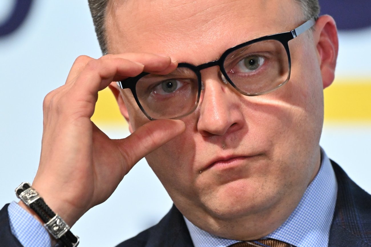 Szymon Hołownia trzymający podtrzymujący oprawkę okularów na twarzy za pomocą prawej dłoni.