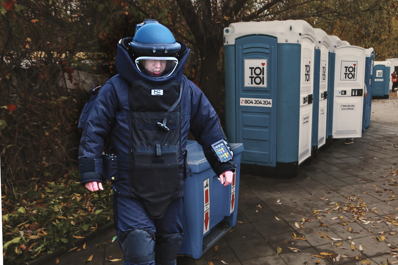 Na zdjęciu policyjny pirotechnik. W tle toaleta toi-toi na cmentarzu.