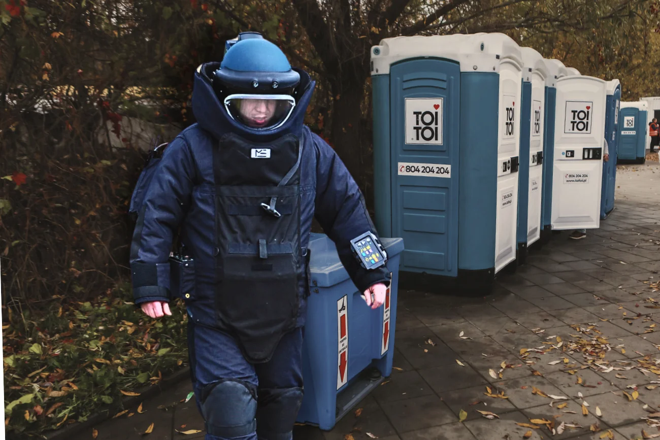Na zdjęciu policyjny pirotechnik. W tle toaleta toi-toi na cmentarzu.