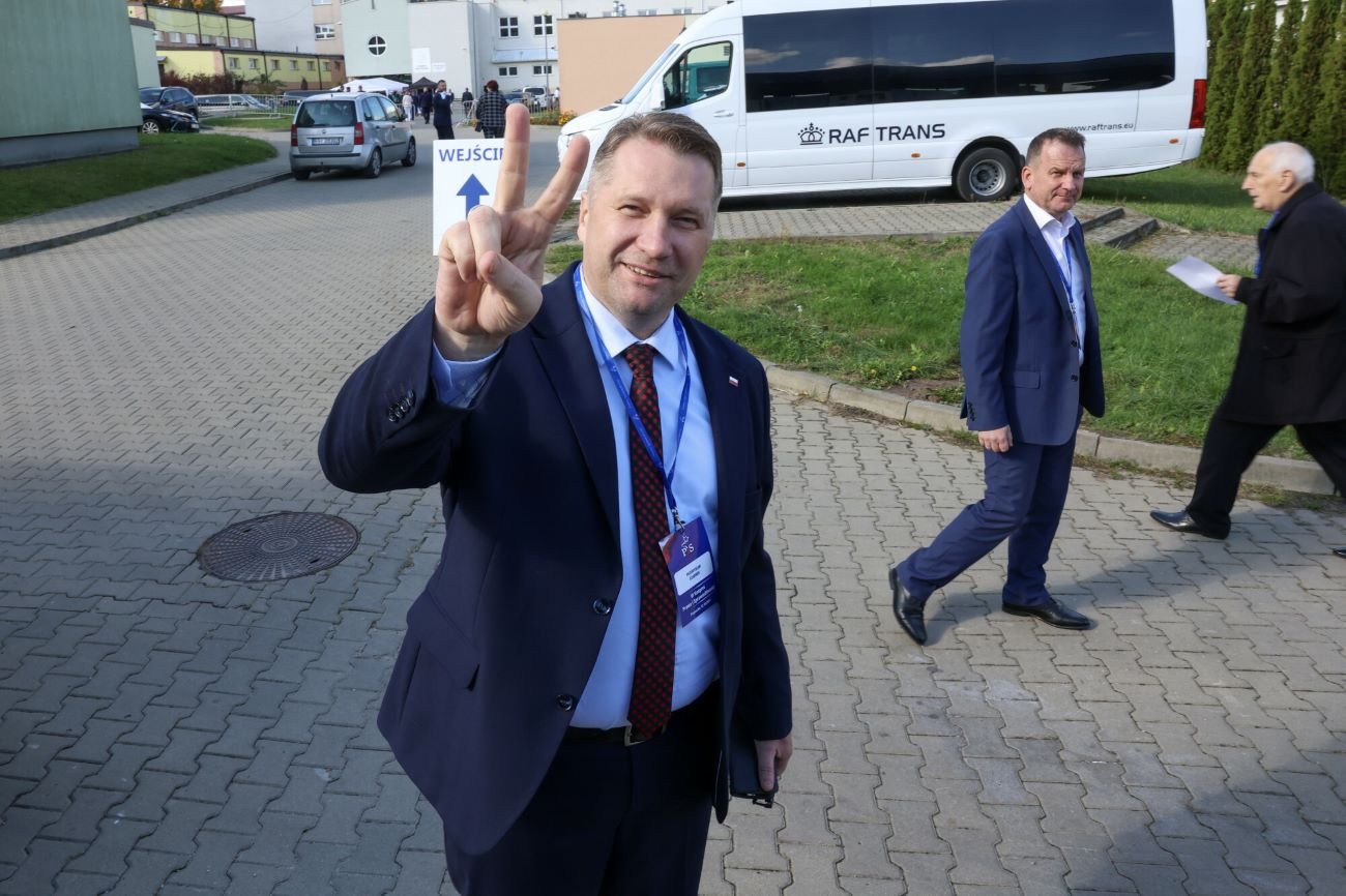 Przemysław Czarnek stojący na ulicy z palcami dłoni ułożonymi w literę V.