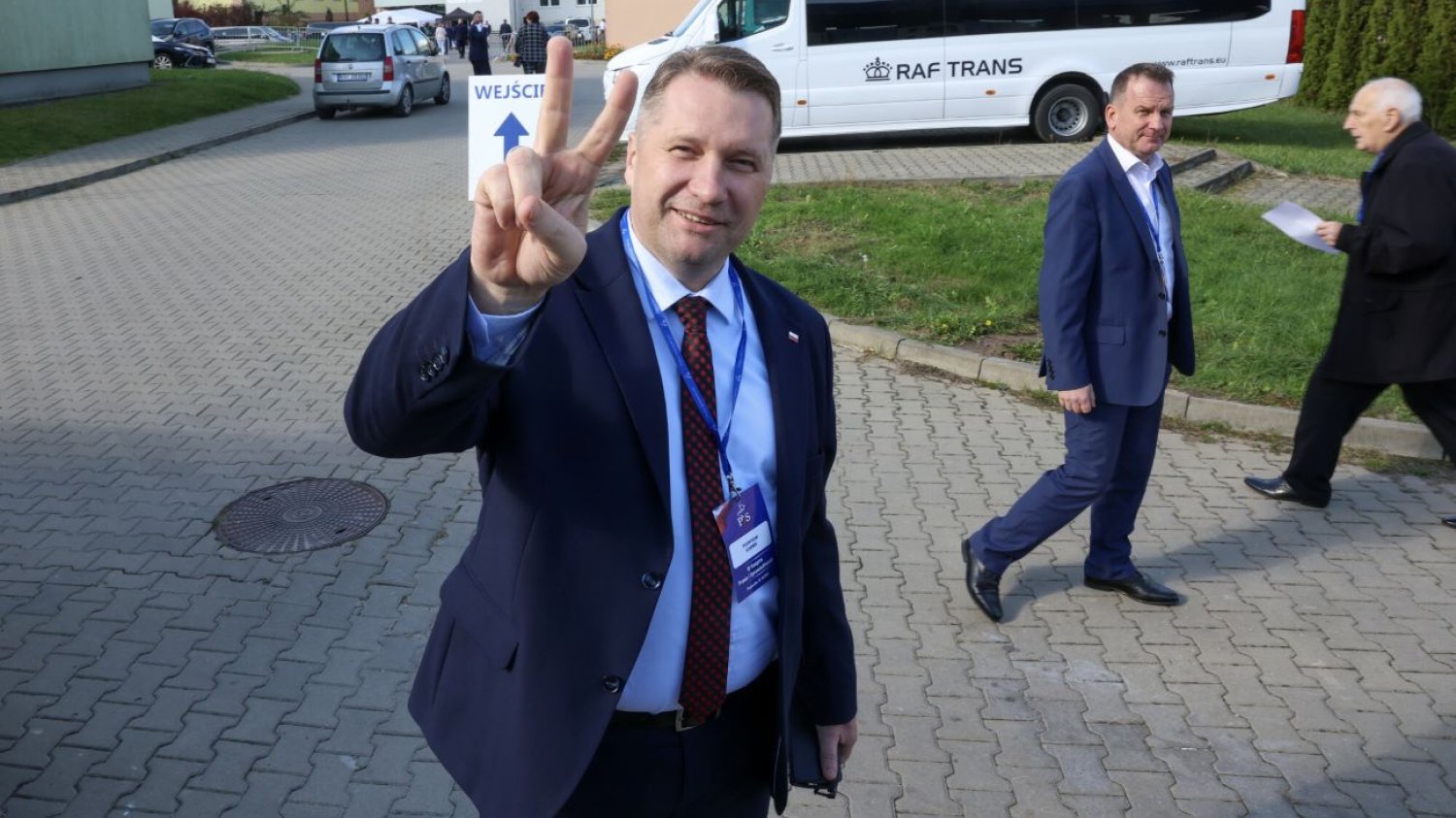 Przemysław Czarnek stojący na ulicy z palcami dłoni ułożonymi w literę V.