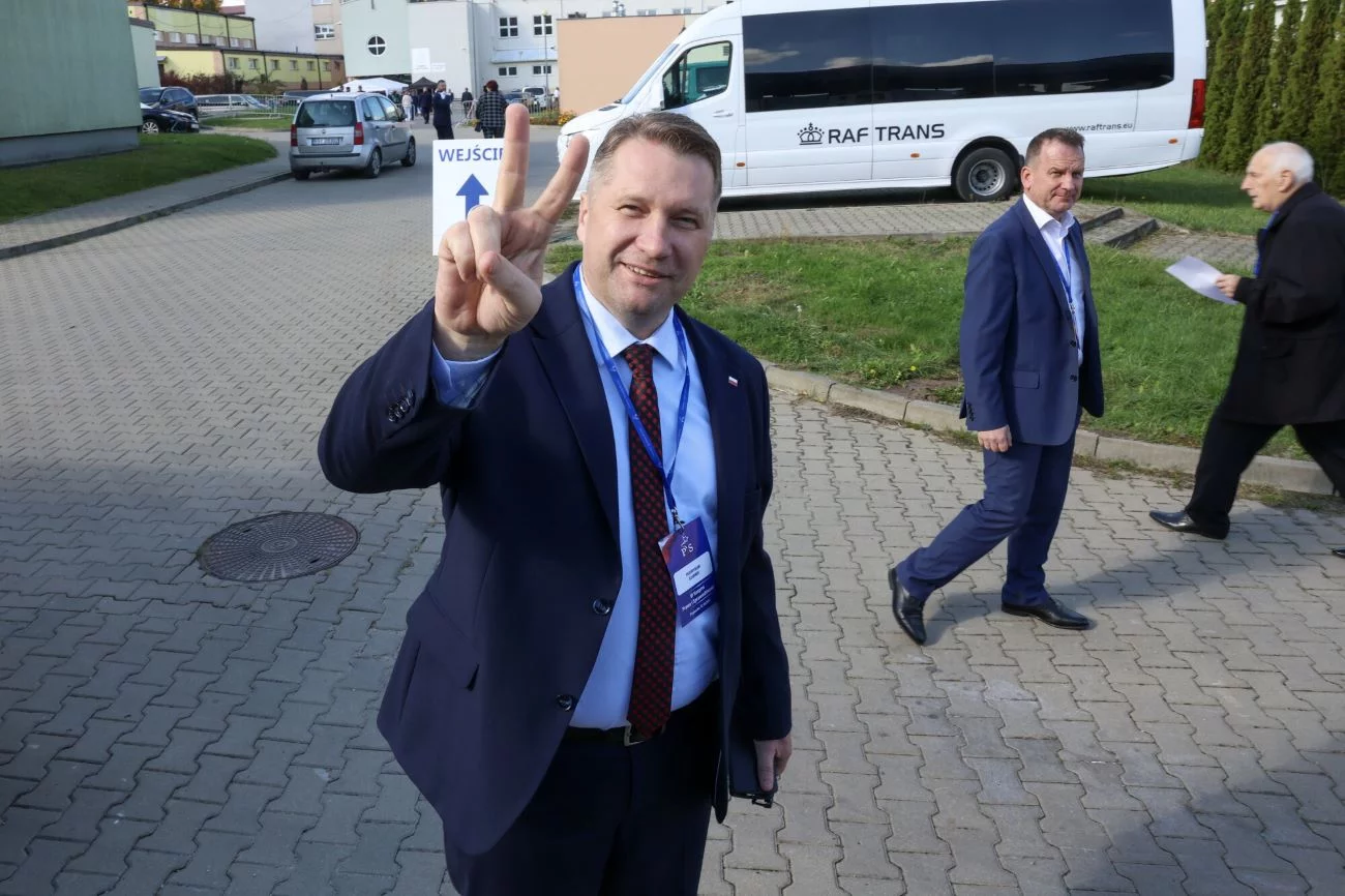 Przemysław Czarnek stojący na ulicy z palcami dłoni ułożonymi w literę V. Przemysław Czarnek stojący na ulicy z palcami dłoni ułożonymi w literę V.