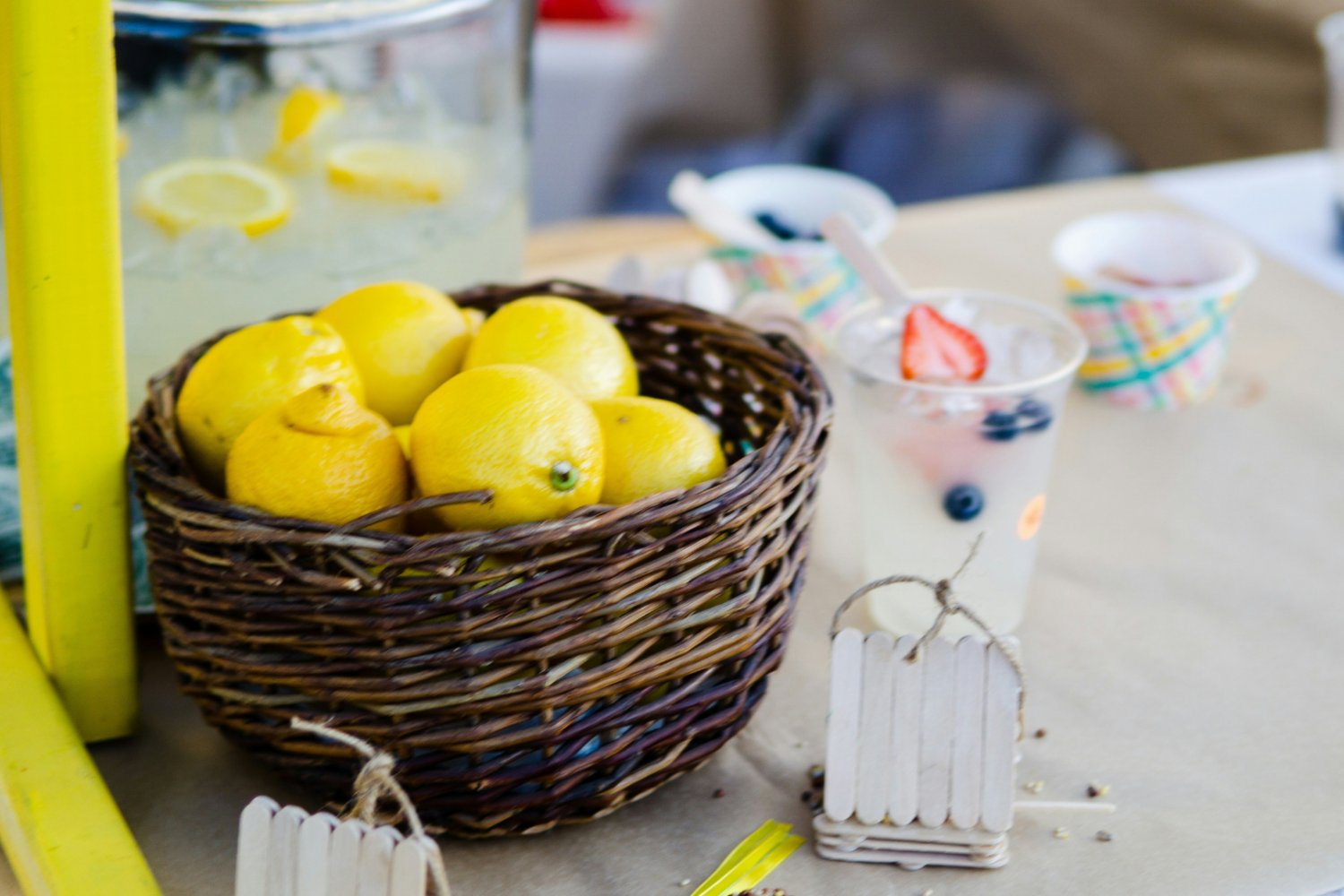 koszyk z cytrynami, szklanki z lemoniadą, naczynie z lemoniadą w tle