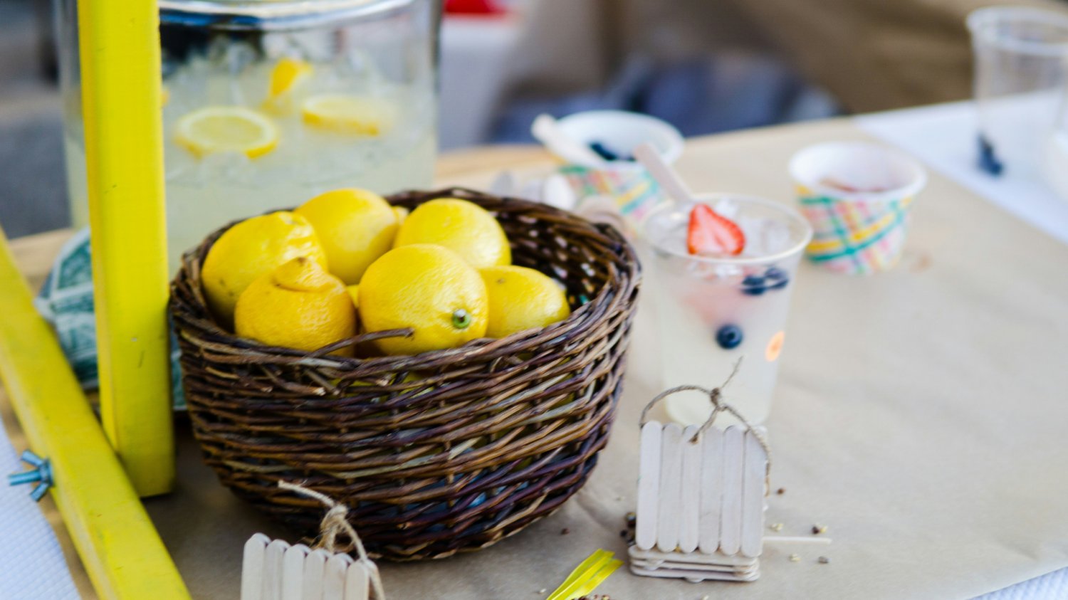 koszyk z cytrynami, szklanki z lemoniadą, naczynie z lemoniadą w tle