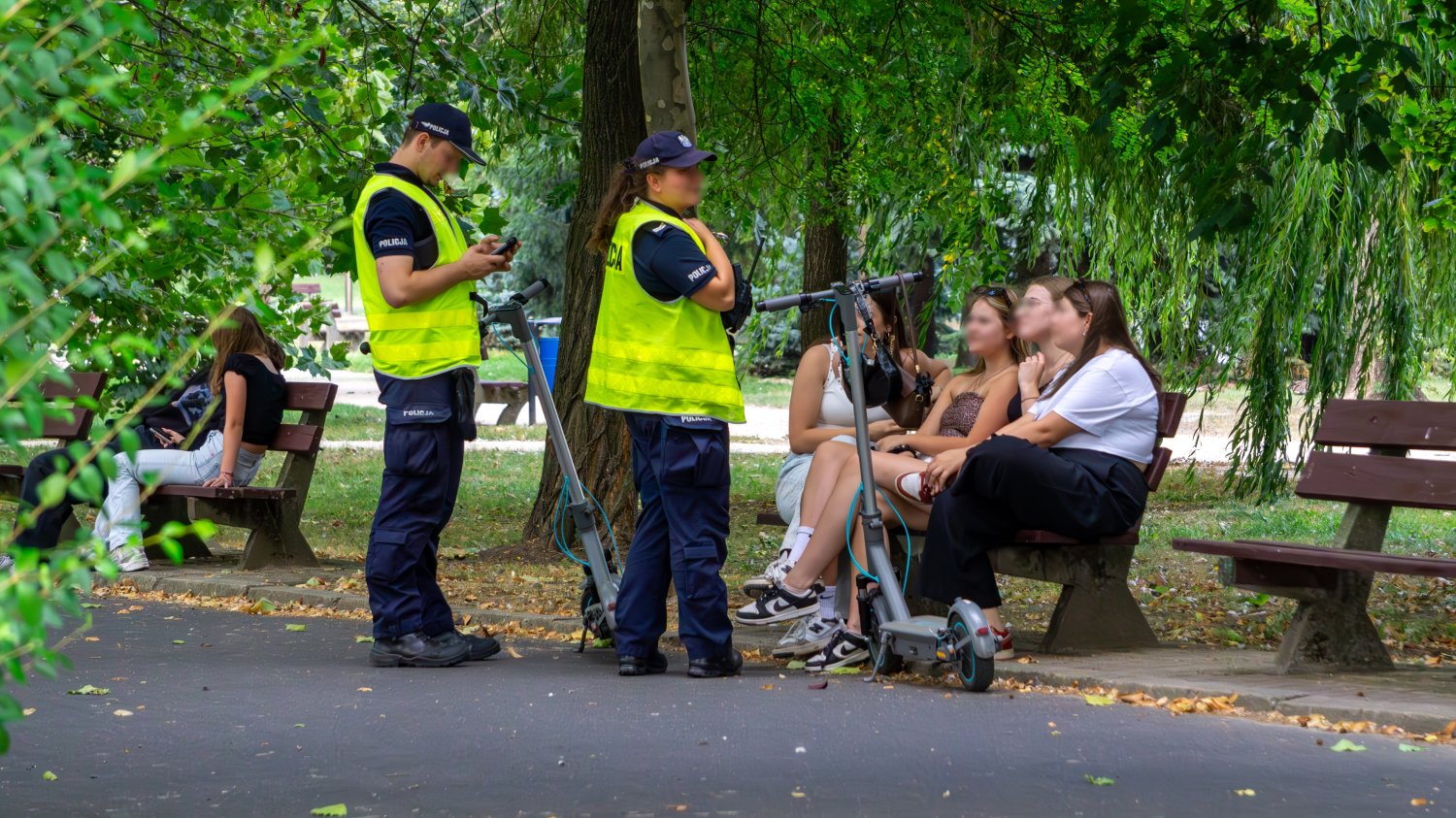 Obowiązkowe kaski dla dzieci i mandat 100 zł od 3 czerwca 2026 r. Policjanci w kamizelkach odblaskowych sprawdzający użytkowników hulajnogi elektrycznej w parku, w pobliżu ławek i zielonych drzew