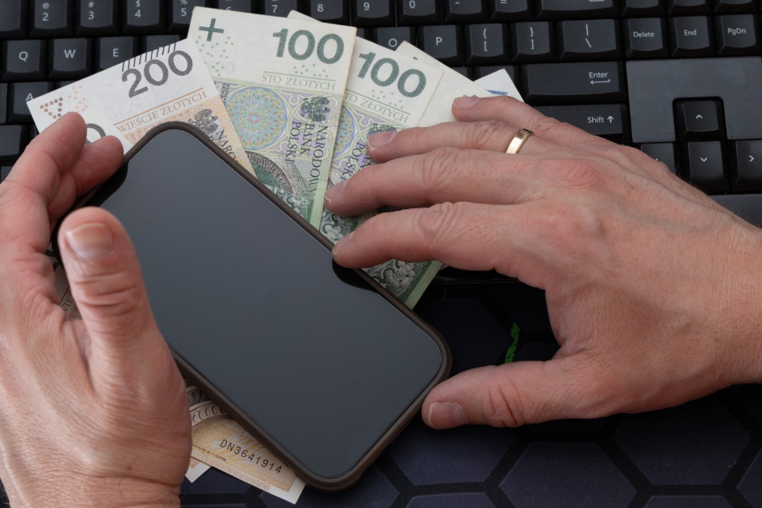Mężczyzna trzyma w ręku telefon z czarnym ekranem, polskie banknoty 200 i 100 zł są ułożone w wentylator na stole, telefon z czarnym ekranem jest umieszczony na rozrzuconych pieniądzach na biurku, wykonywany jest przelew online
