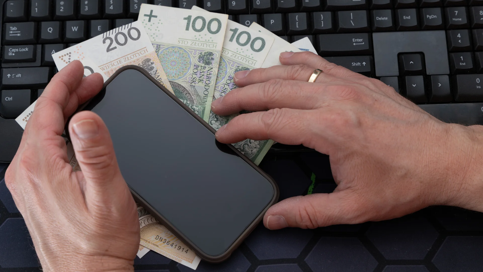 Ryzykowne przelewy 2026. Co alarmuje bank i skarbówkę? Mężczyzna trzyma w ręku telefon z czarnym ekranem, polskie banknoty 200 i 100 zł są ułożone w wentylator na stole, telefon z czarnym ekranem jest umieszczony na rozrzuconych pieniądzach na biurku, wykonywany jest przelew online