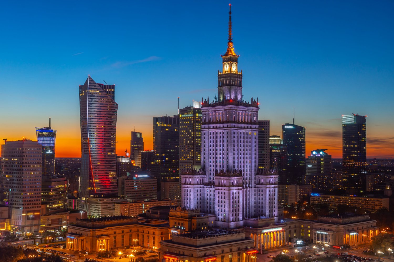 Widok z lotu ptaka na panoramę Warszawy o zmierzchu, z oświetlonym Pałacem Kultury i Nauki.