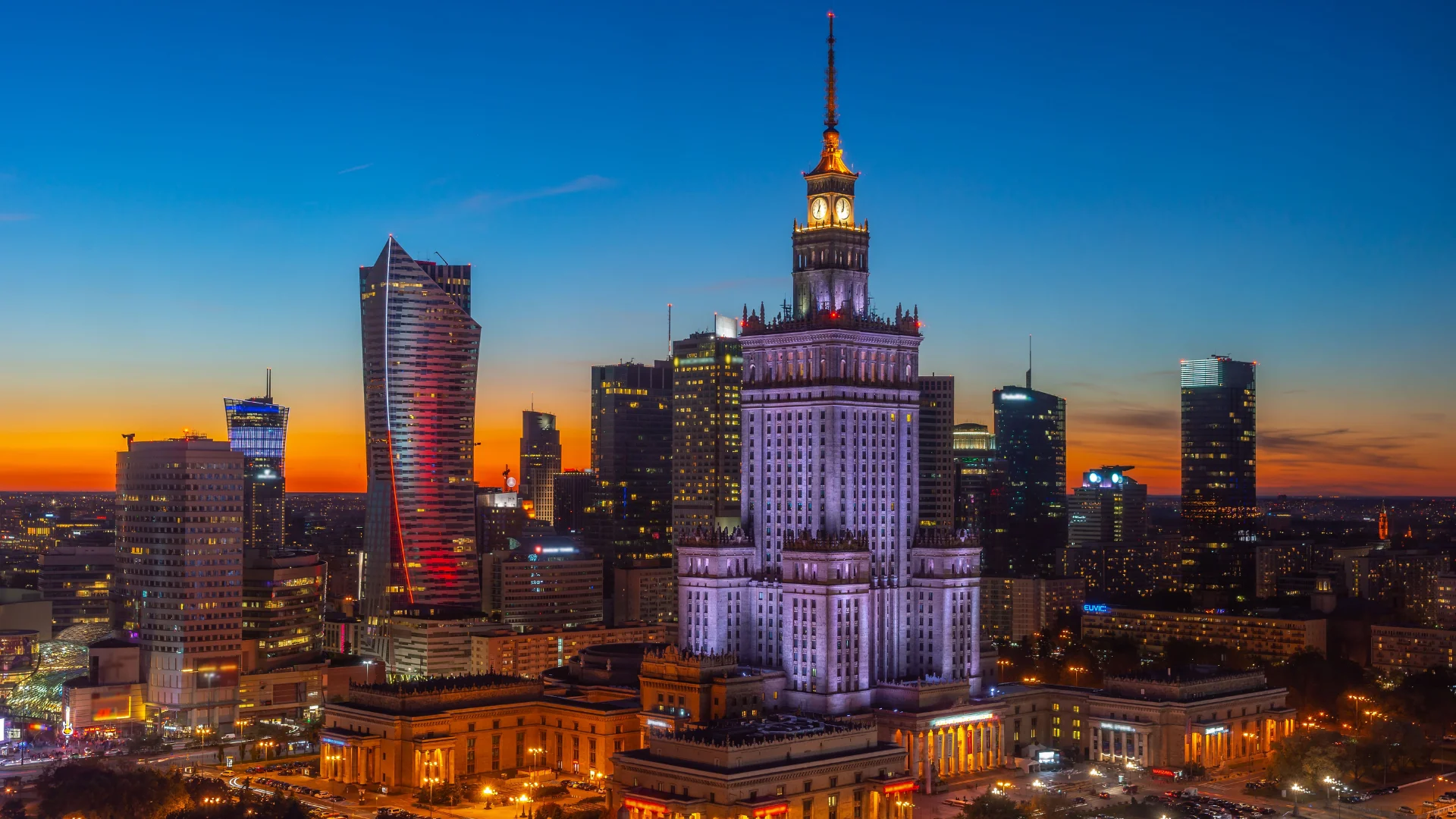 Widok z lotu ptaka na panoramę Warszawy o zmierzchu, z oświetlonym Pałacem Kultury i Nauki.