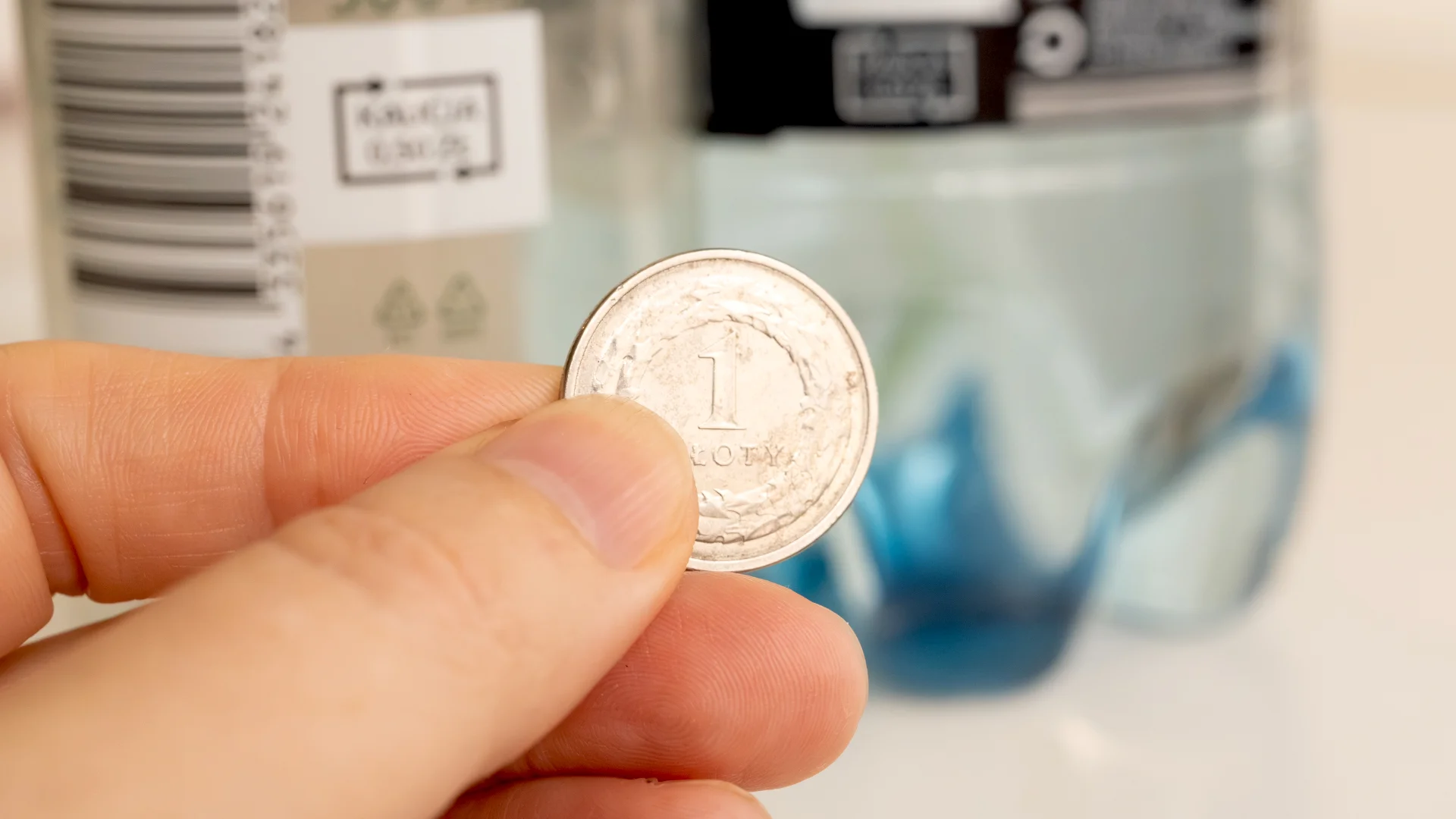 Zbliżenie na dłoń trzymającą złotówkę, a w tle butelki z kaucją